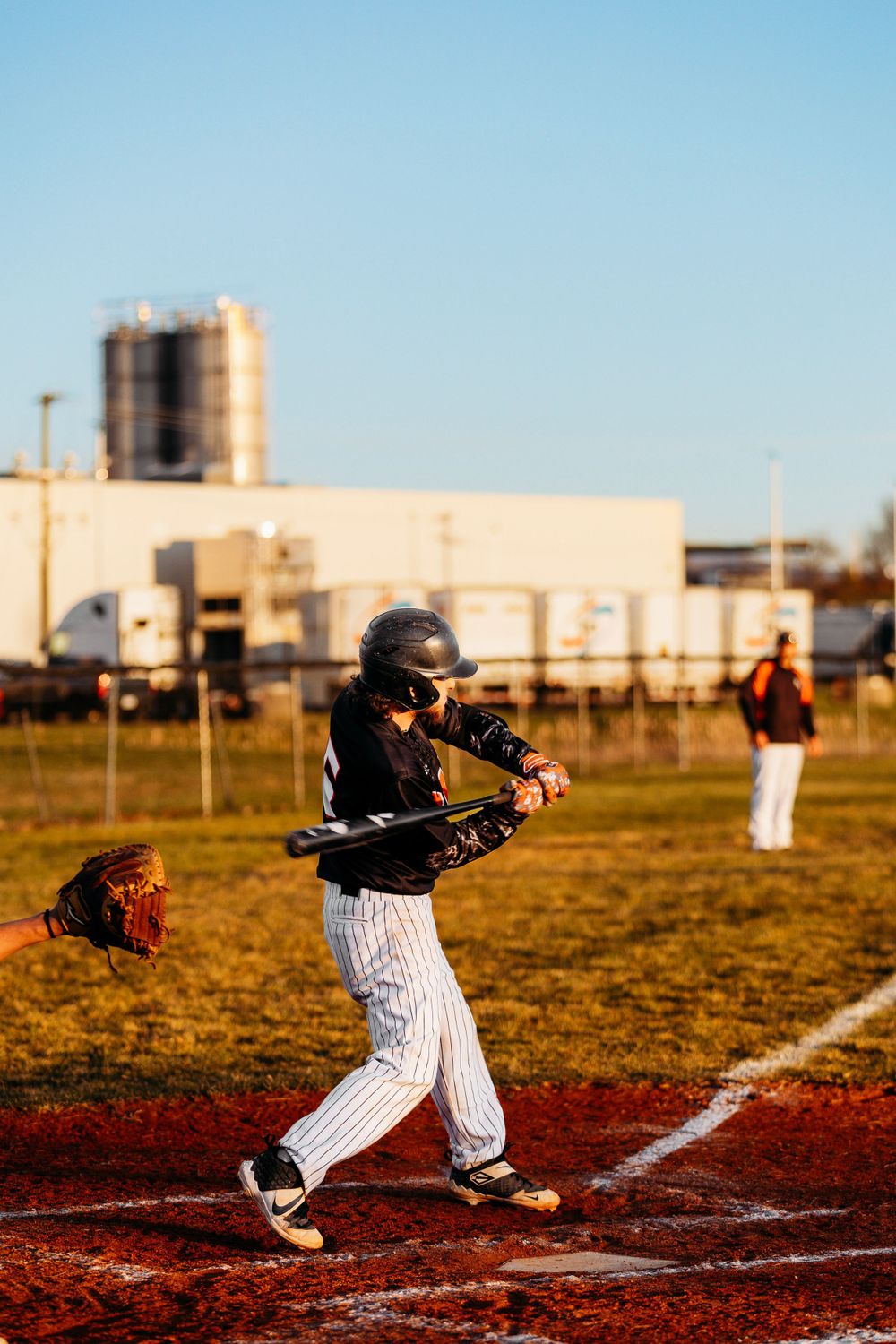Youth baseball batter in black pinstripe uniform swings at a pitch during a golden hour game.
