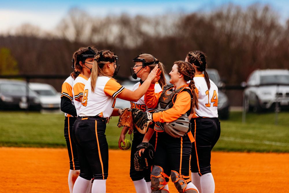Female softball team in orange and black uniforms huddles together celebrating on the field.