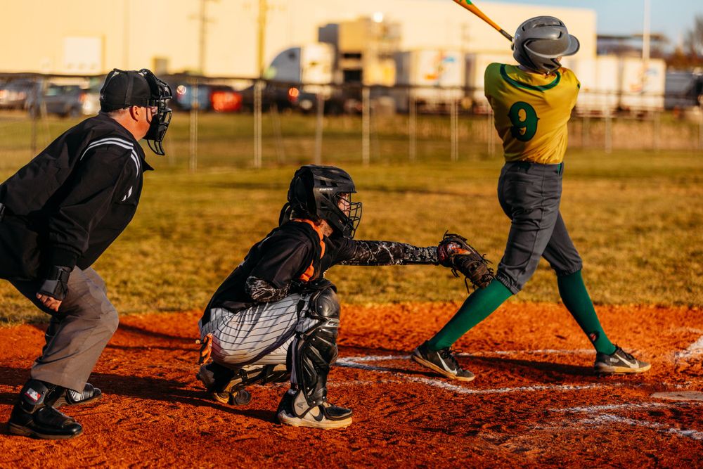 Baseball batter in green jersey swings as catcher and umpire watch during outdoor youth game.