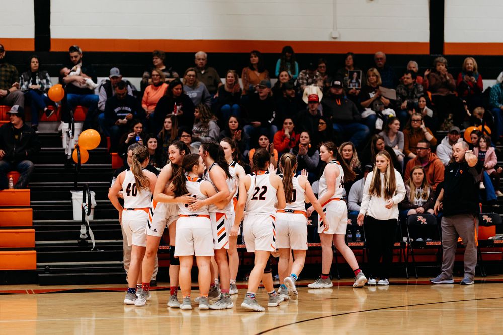 Girls high school basketball team huddles together on court before a game with a packed crowd.
