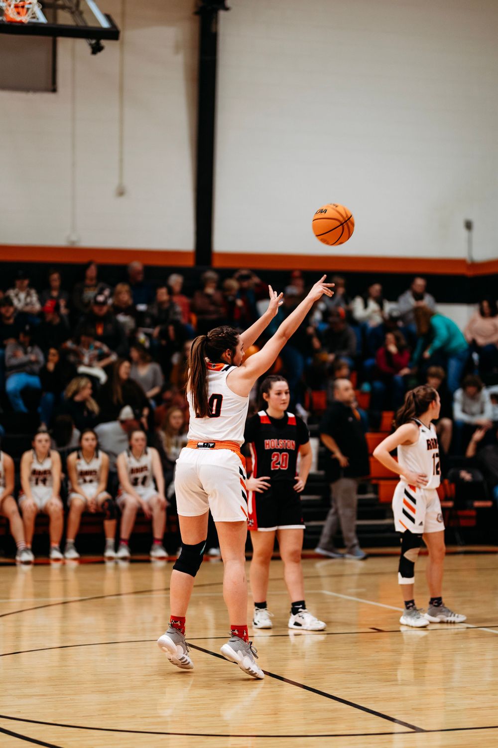 Girls basketball player releases a jump shot while opponents and teammates watch during a high school game.
