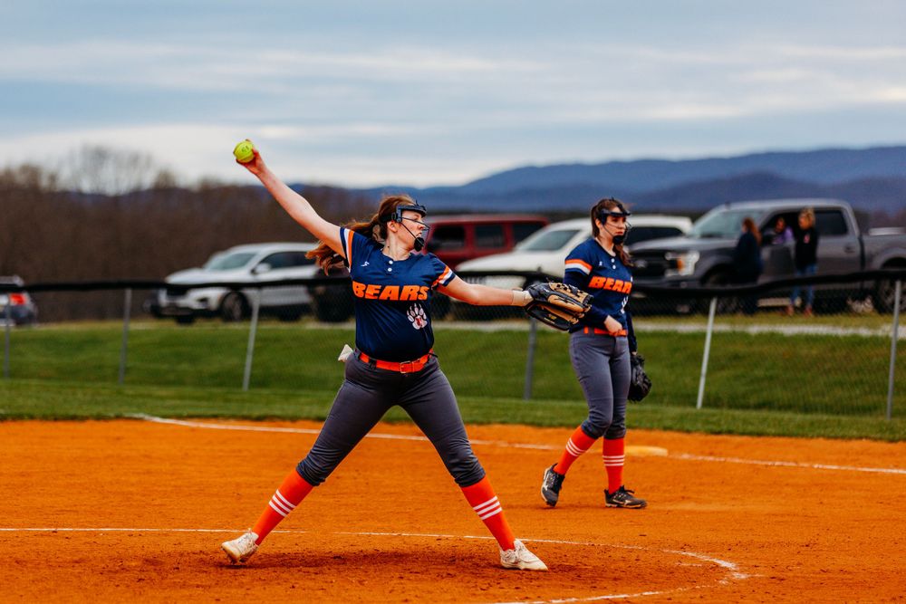 Softball pitcher in orange Bears uniform winds up to deliver a pitch during a game on red dirt field.