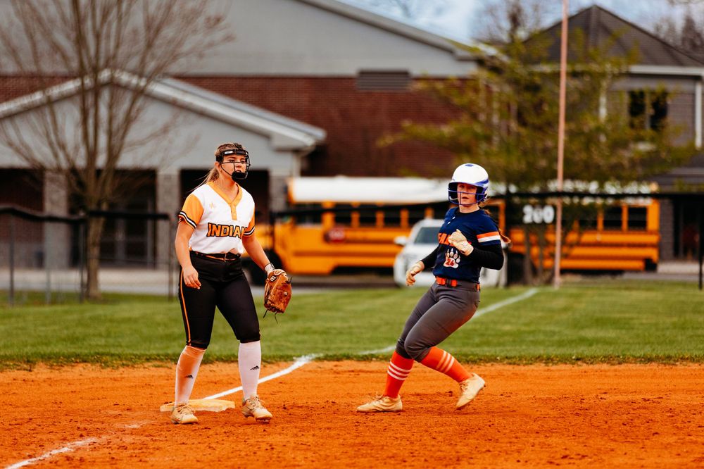Two softball players near first base with a yellow school bus visible in the background.
