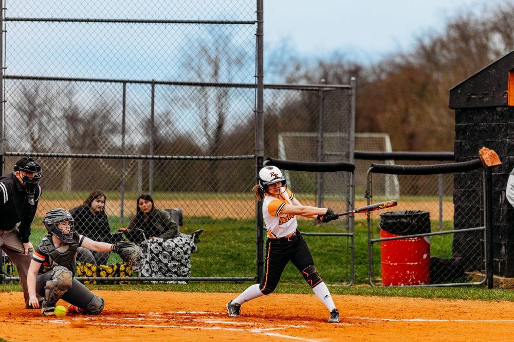 Softball player in orange uniform swinging bat at home plate during a game on a red dirt field.