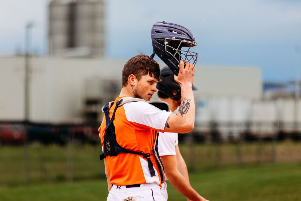 Baseball catcher in orange and black gear removing helmet on an outdoor field.