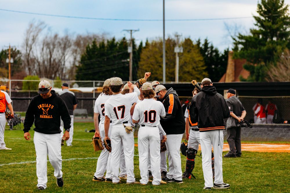 High school baseball team huddles together on green field wearing white and orange uniforms during game.