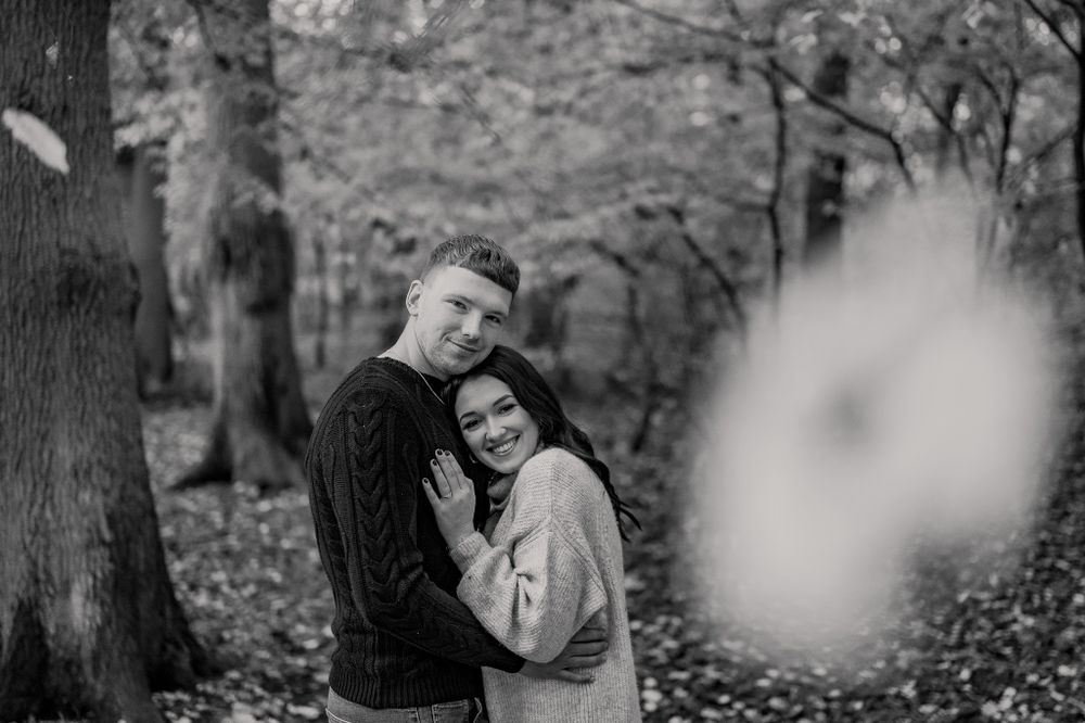 Black and white photo of a tender embrace between a couple in a wooded setting with fallen leaves.