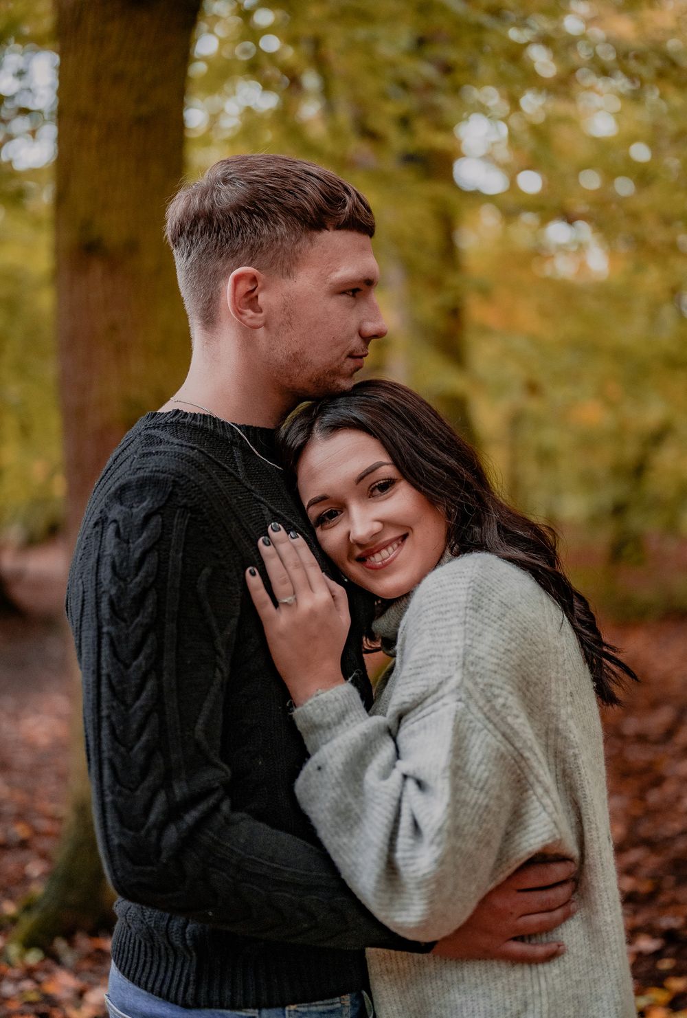 A couple in cozy sweaters sharing an intimate moment in a fall forest setting with warm lighting.