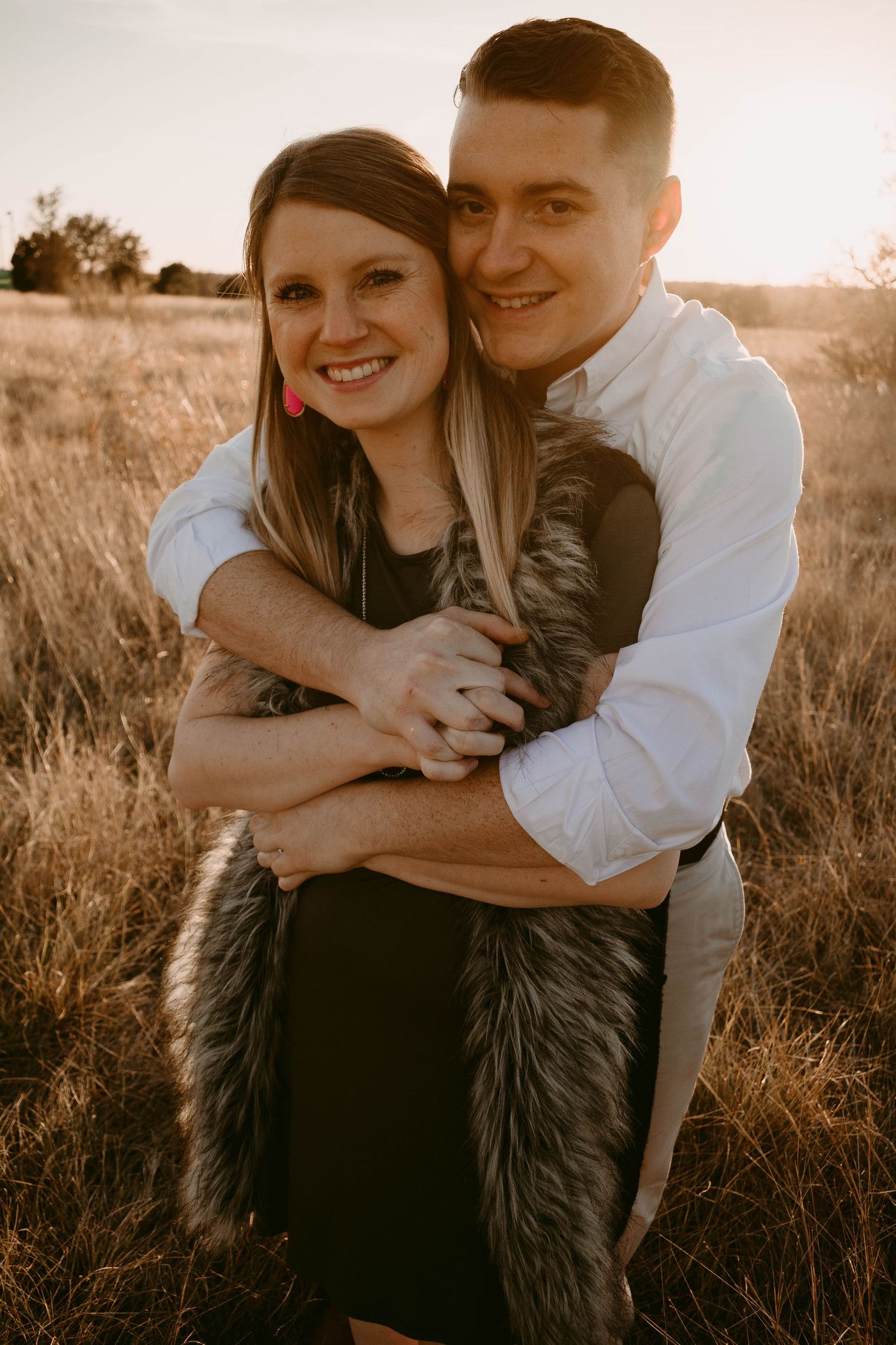 Couple wearing black dress and white shirt embracing while wrapped in fur vest outdoors.