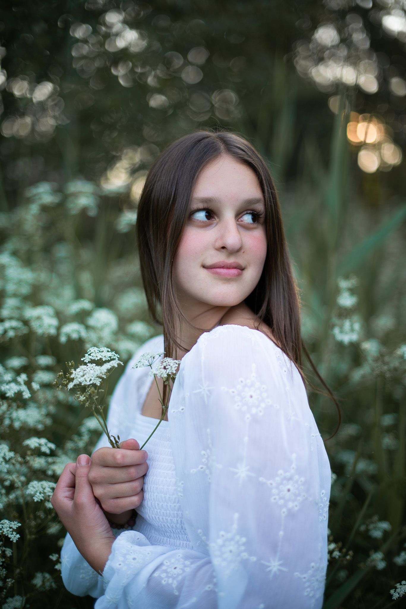 Girl in white dress holding delicate flowers against moody forest backdrop.
