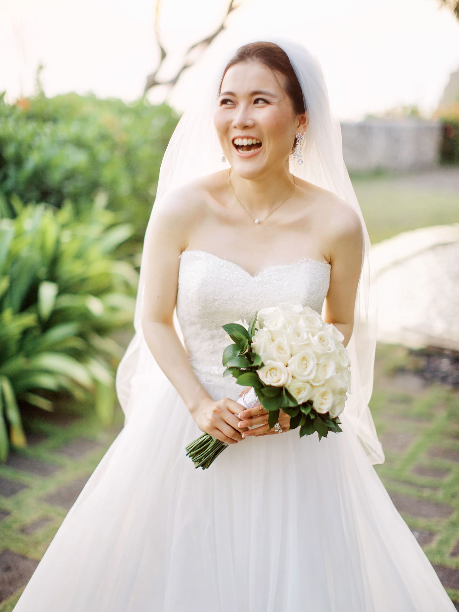 A luxury bride in a white dress radiantly smiles, clutching her bouquet at Bulgari Resort Bali.