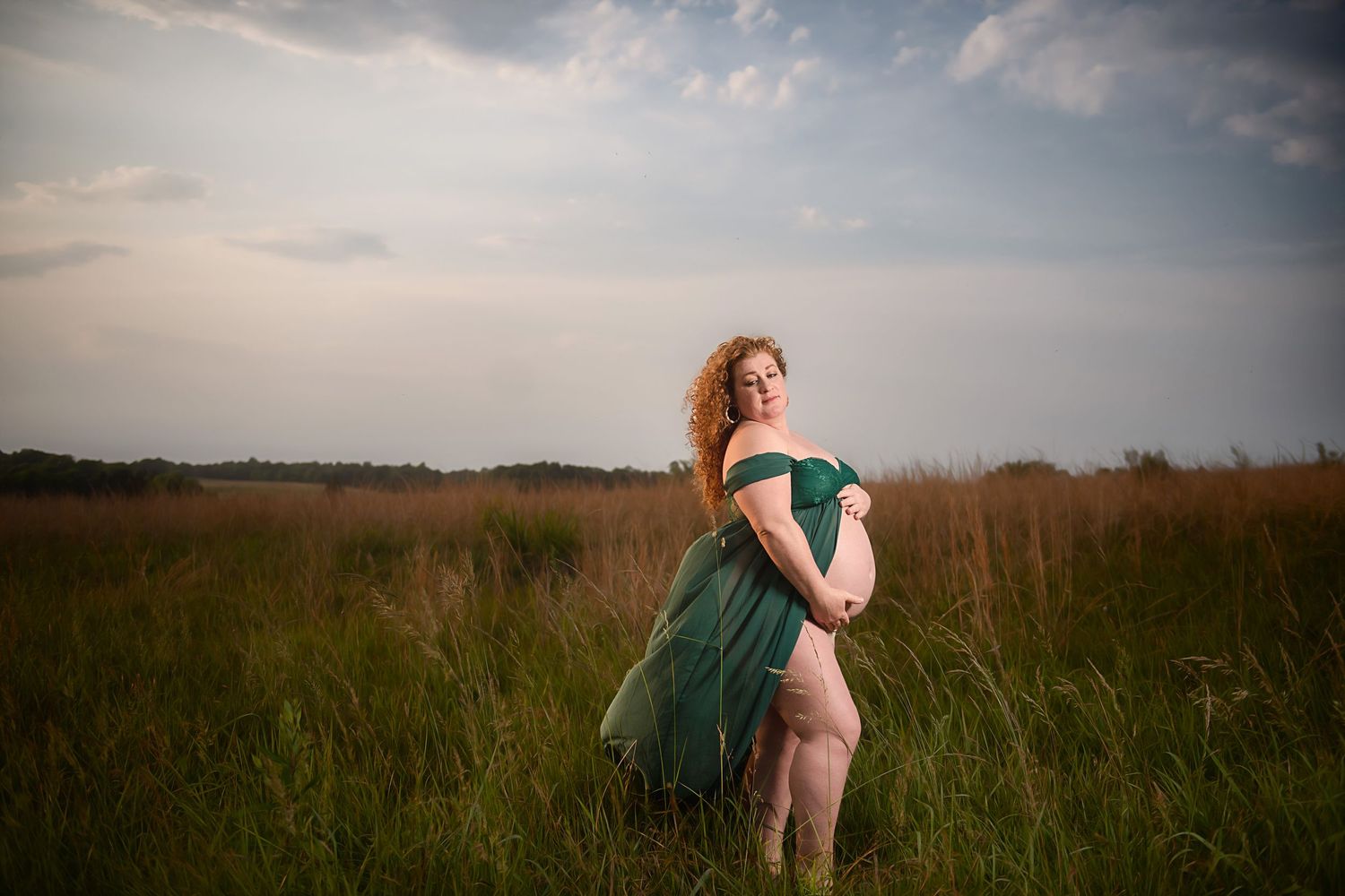 Maternity portrait of woman in emerald dress standing in golden field at sunset.