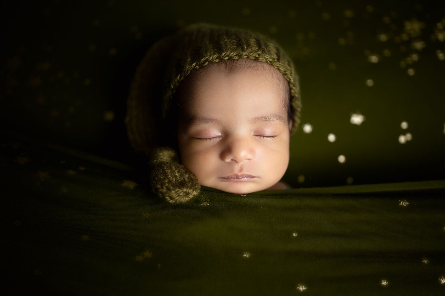 Sleeping newborn in green knit wrap with delicate lace headband against dark background.