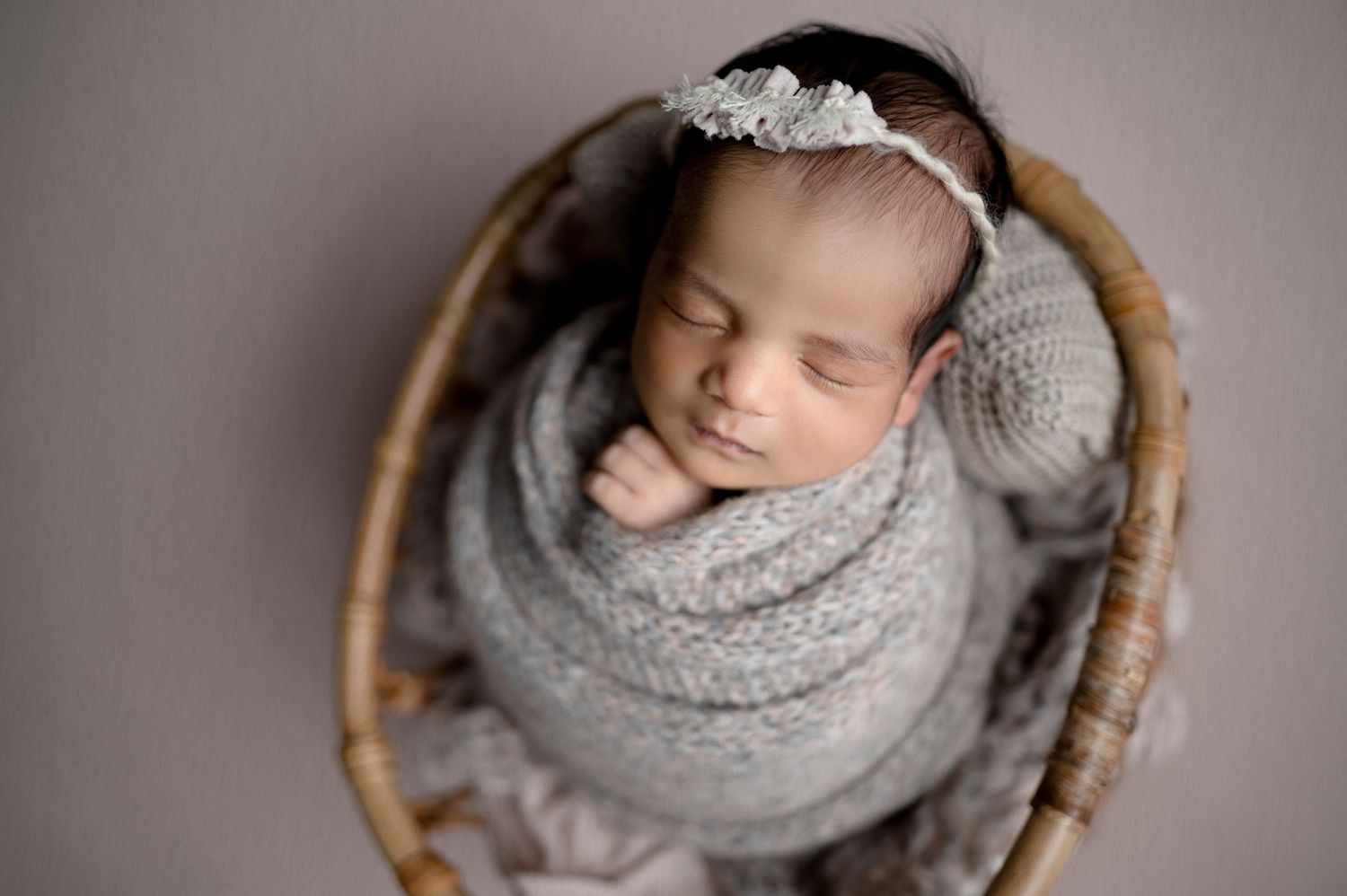 Newborn baby sleeping in grey knit wrap inside rustic wooden basket.