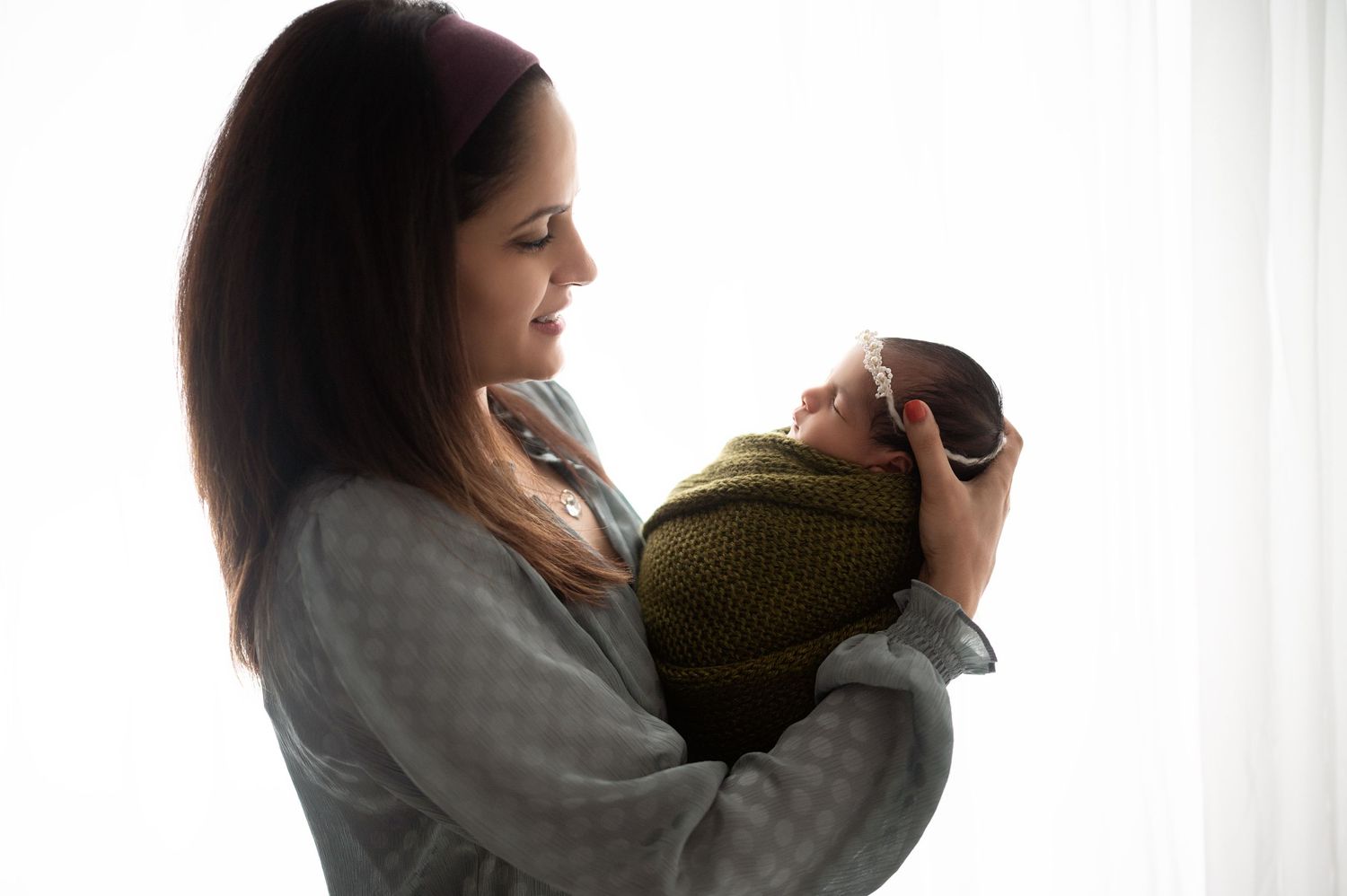 Mother in grey sweater holding swaddled newborn in silhouette against window light.