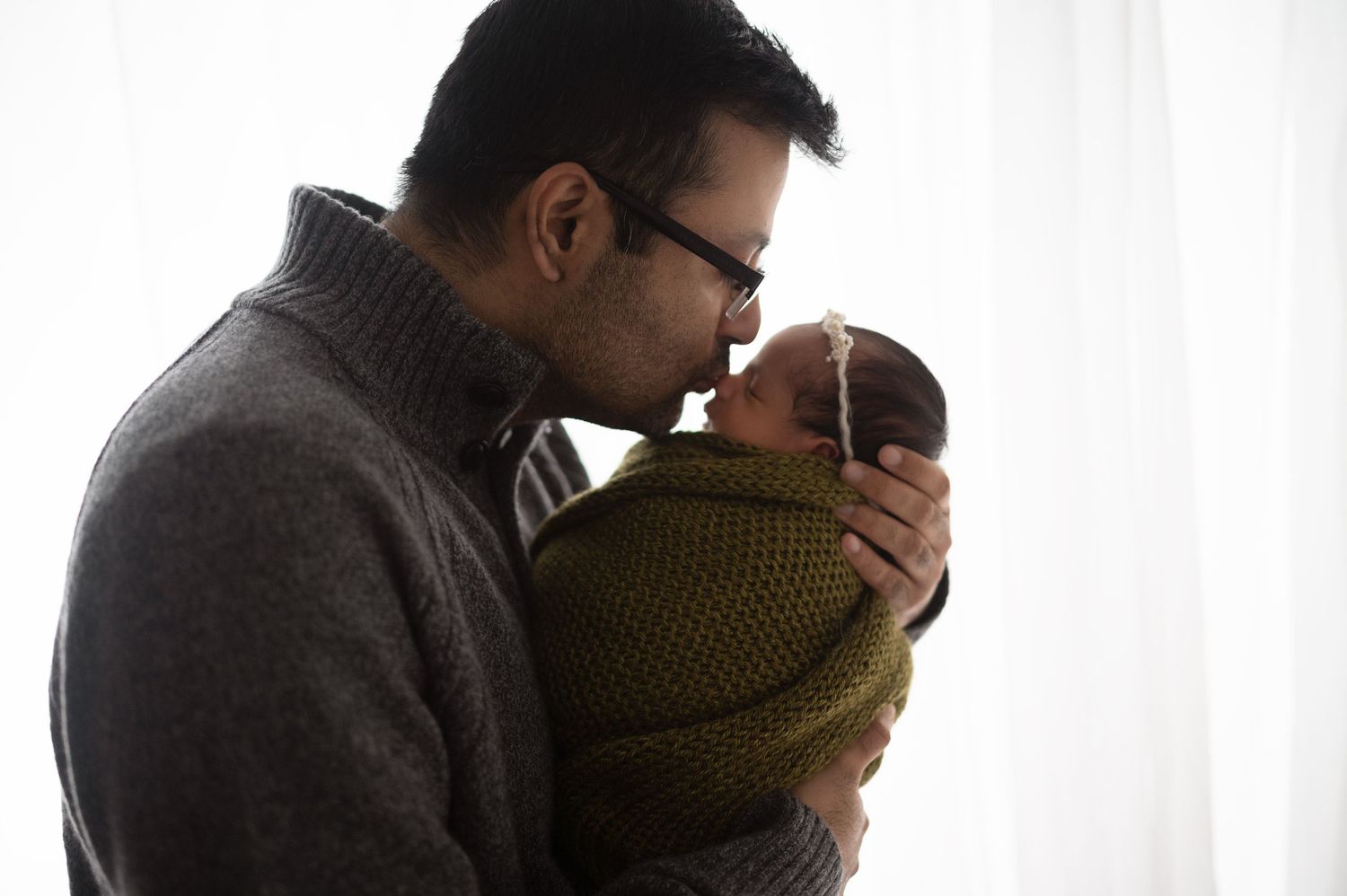 A parent cradles a small baby wrapped in olive green knit wear by a bright window.