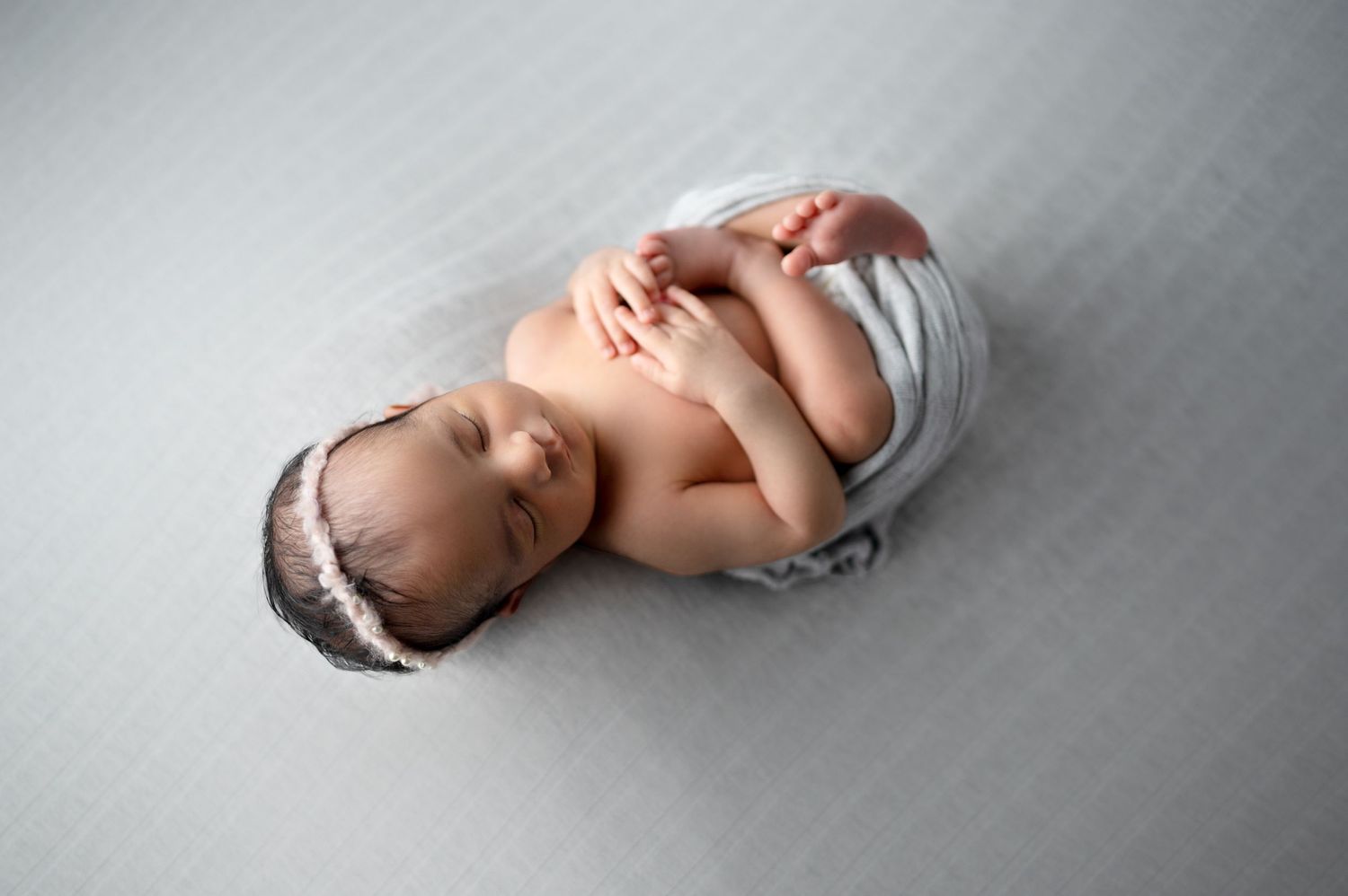 Sleeping newborn baby wrapped in white fabric lying on grey backdrop during professional photoshoot.