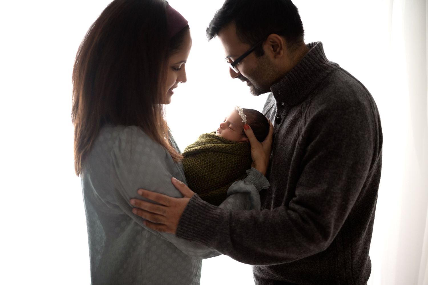 Parents tenderly holding their swaddled newborn in silhouette against bright window.