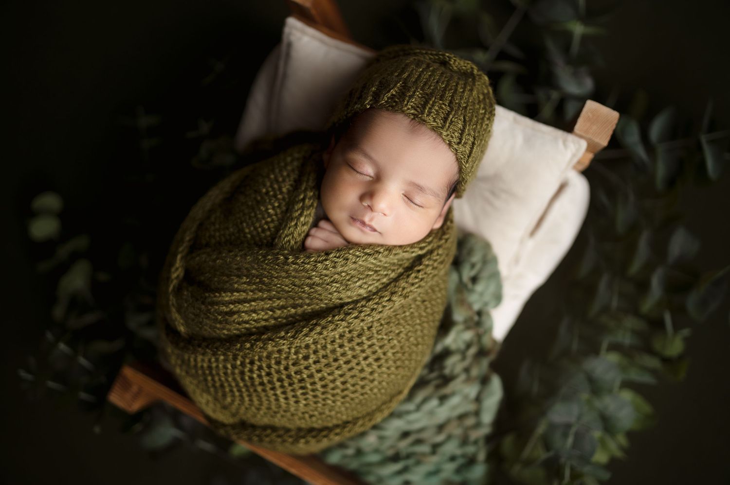 Sleeping infant wrapped in olive green knit blanket with foliage backdrop.
