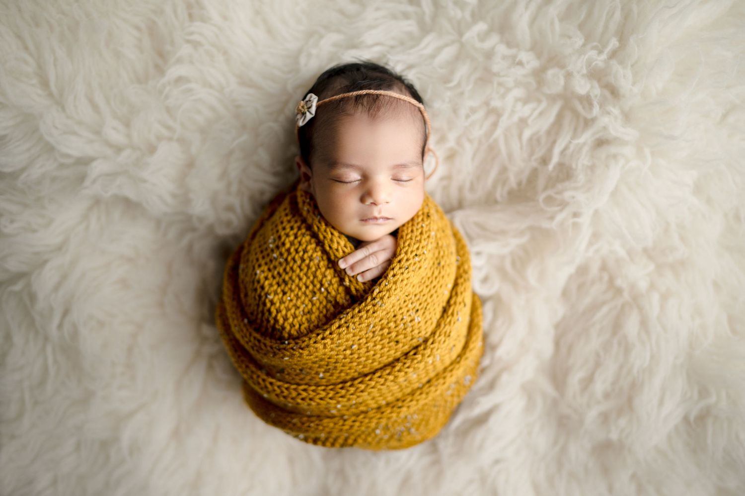 Sleeping newborn wrapped in mustard yellow knit blanket on white fluffy backdrop.