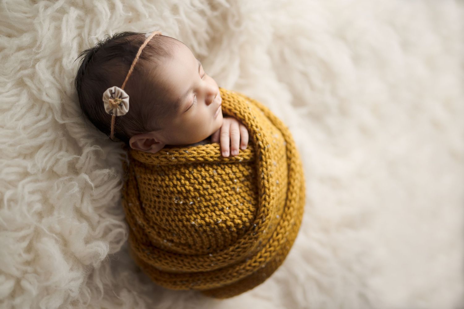 Artistic newborn photos showing baby wrapped in golden knit blanket on cream textured surface.