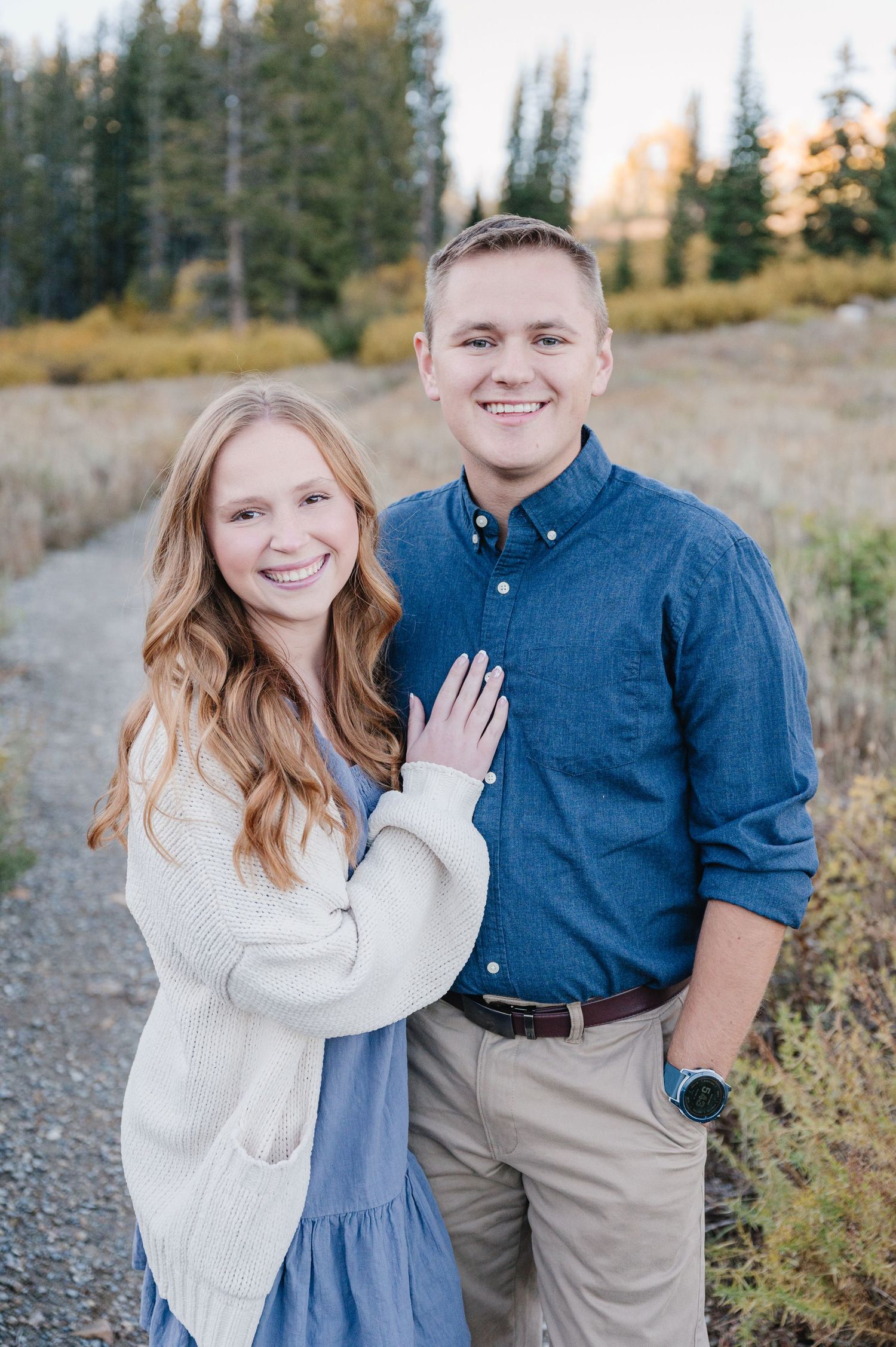 Couple posing for engagement photos on a mountain trail surrounded by pine trees.