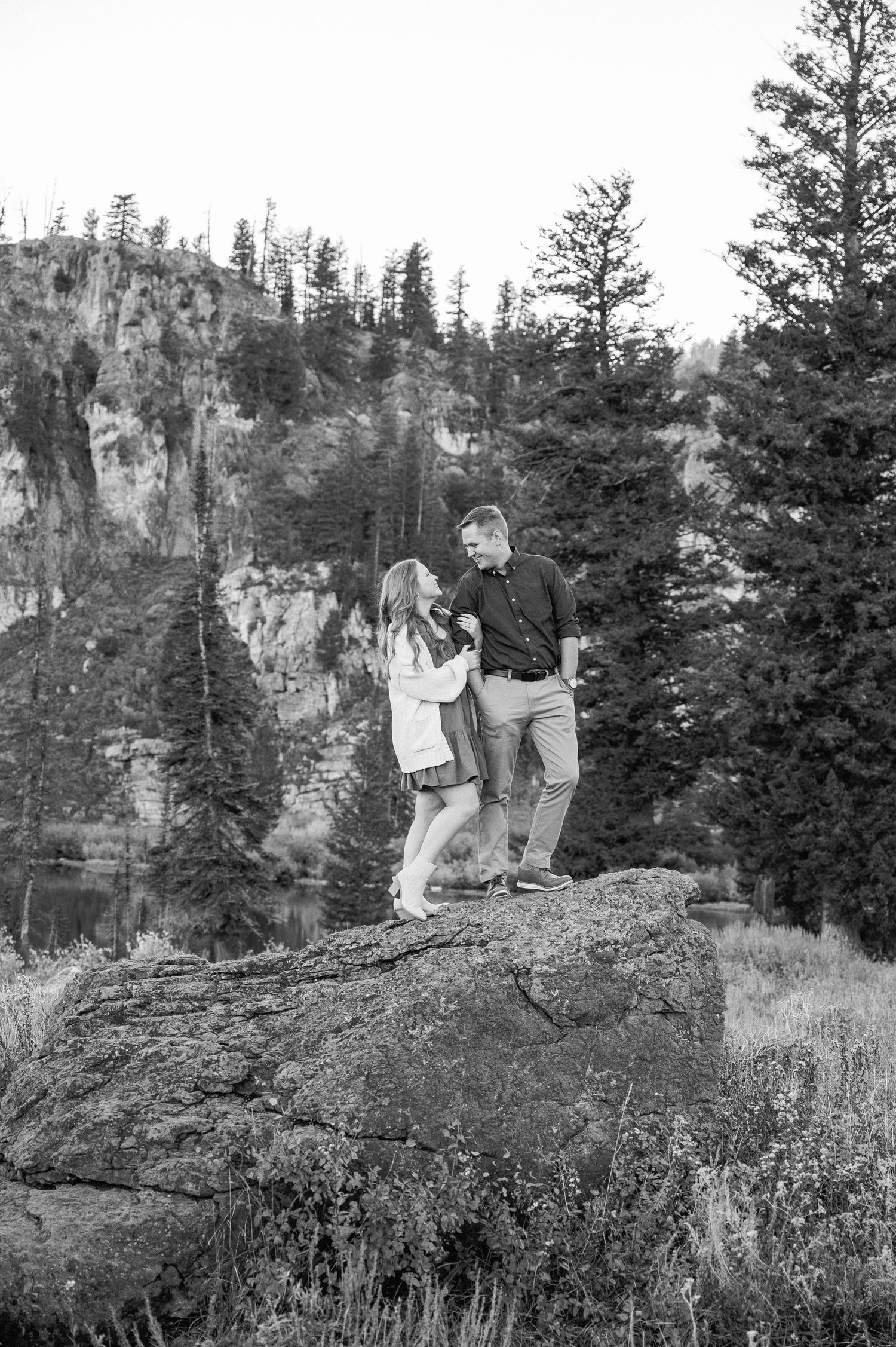 Couple standing on rocky outcrop with mountain cliffs and forest in black and white.