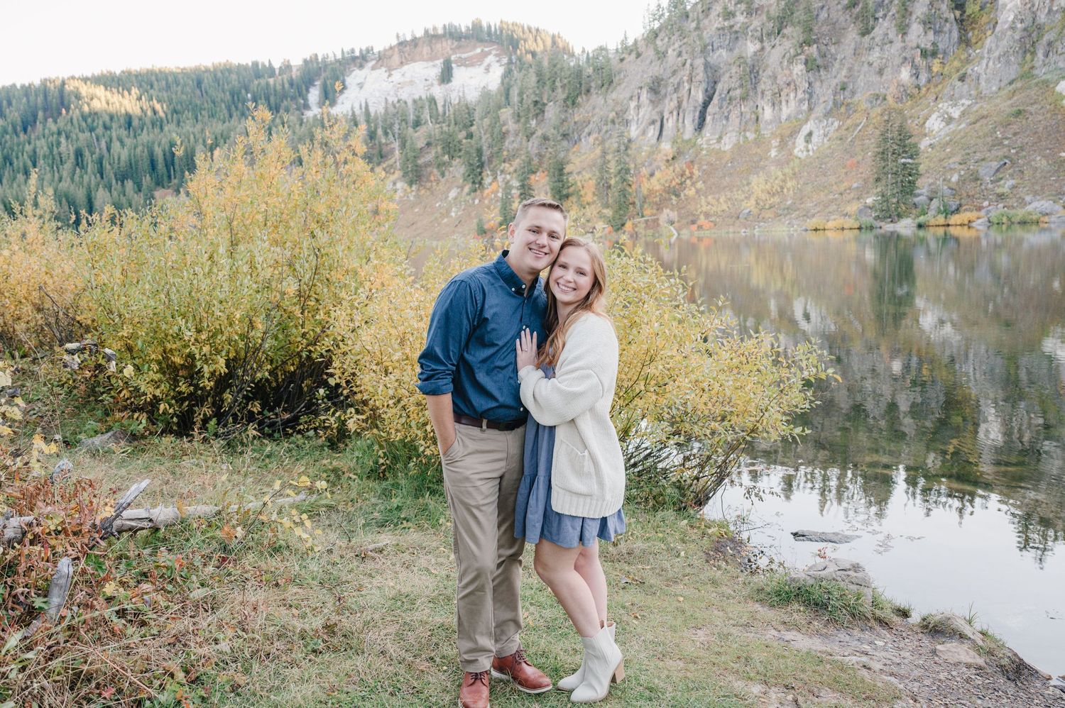 Romantic couple posing by a serene mountain lake with cliffs in the background.