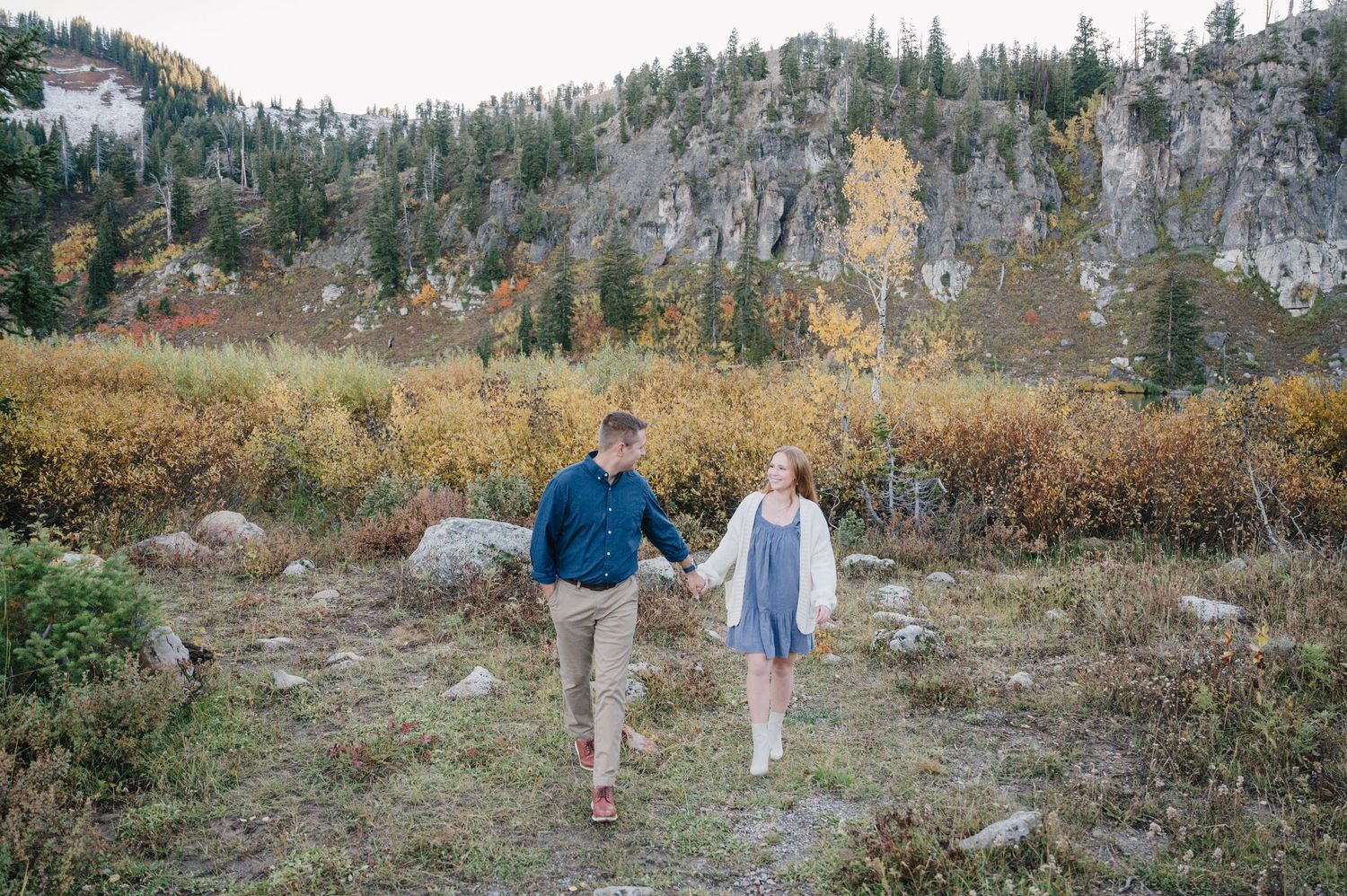 Couple walking hand in hand through a mountain valley with evergreen trees.