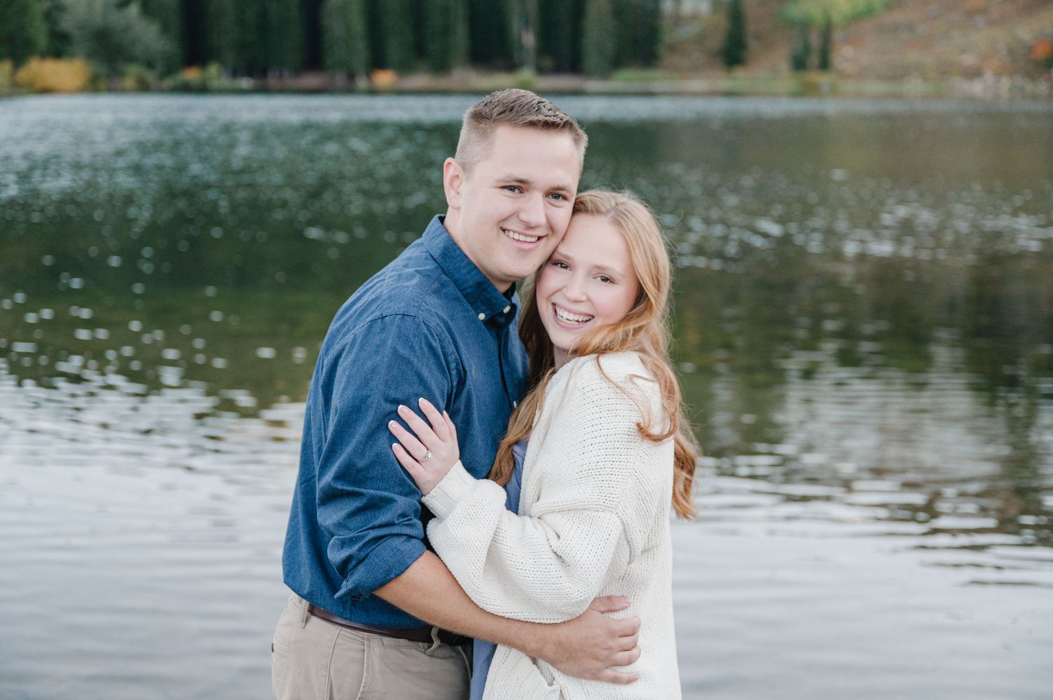 Couple in casual attire embracing by a tranquil lake with mountains in the background.