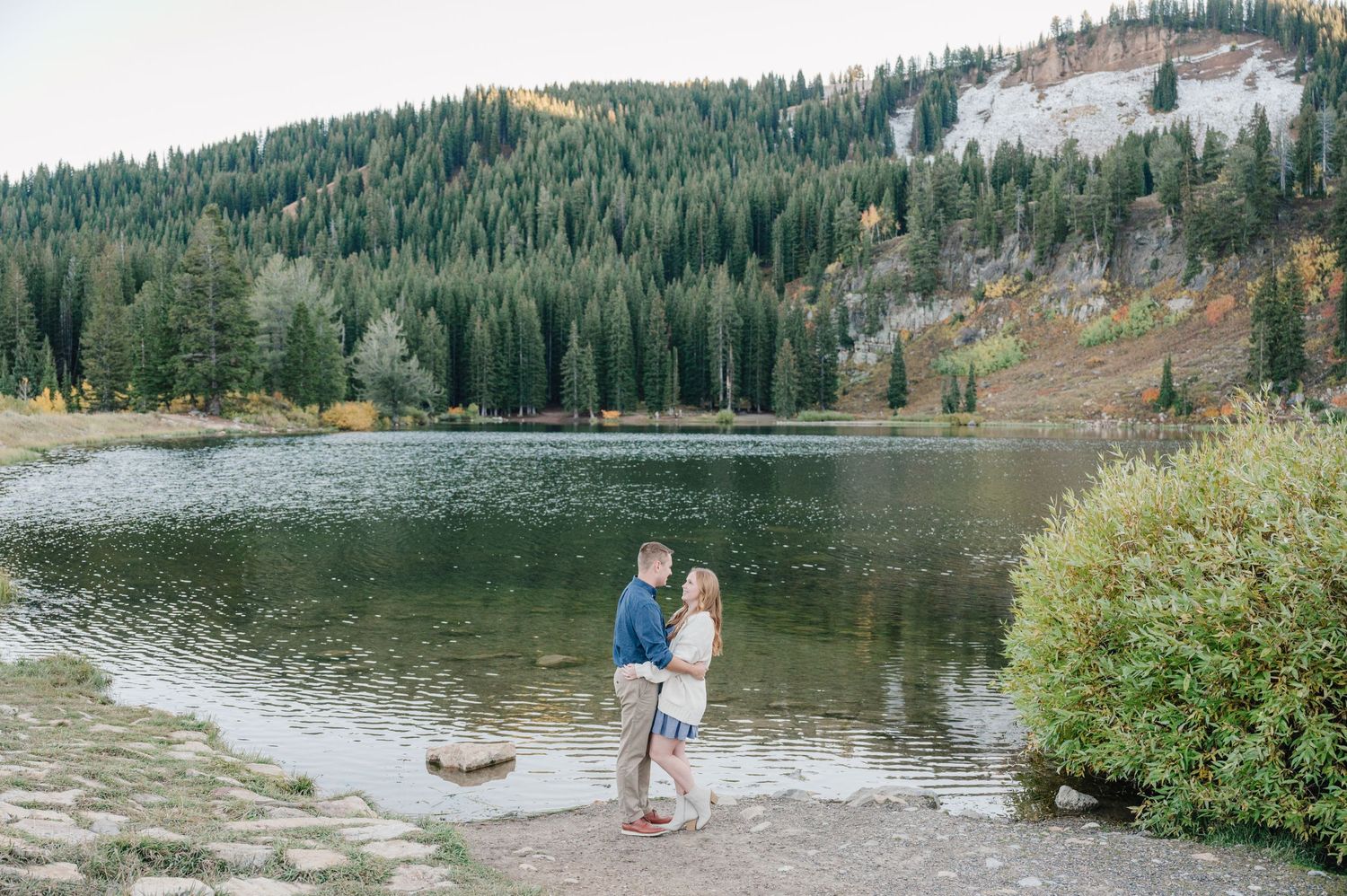 Couple standing together at the edge of a mountain lake surrounded by pine forests.