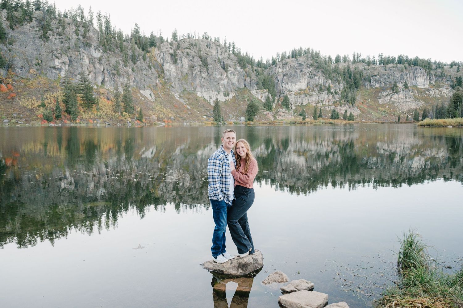 Couple embracing on rocks by reflective mountain lake during autumn.