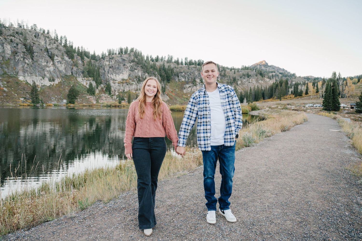 A couple walks hand in hand along a mountain lake trail with pine trees and mountains in the background.