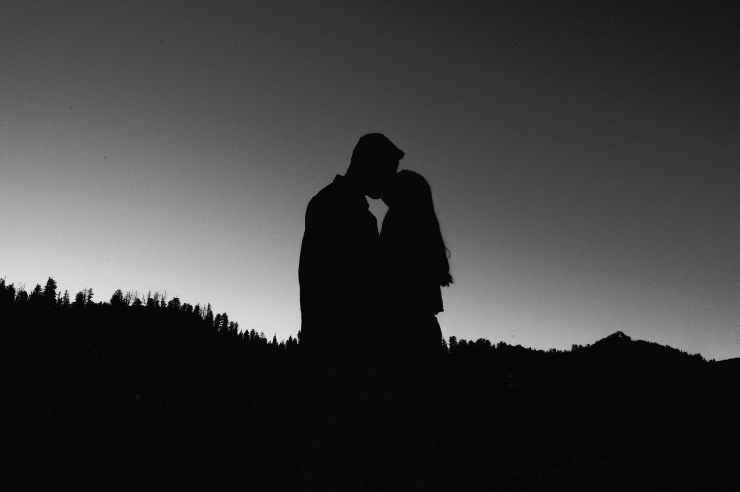 Silhouetted couple against sunset sky with forest treeline in background.