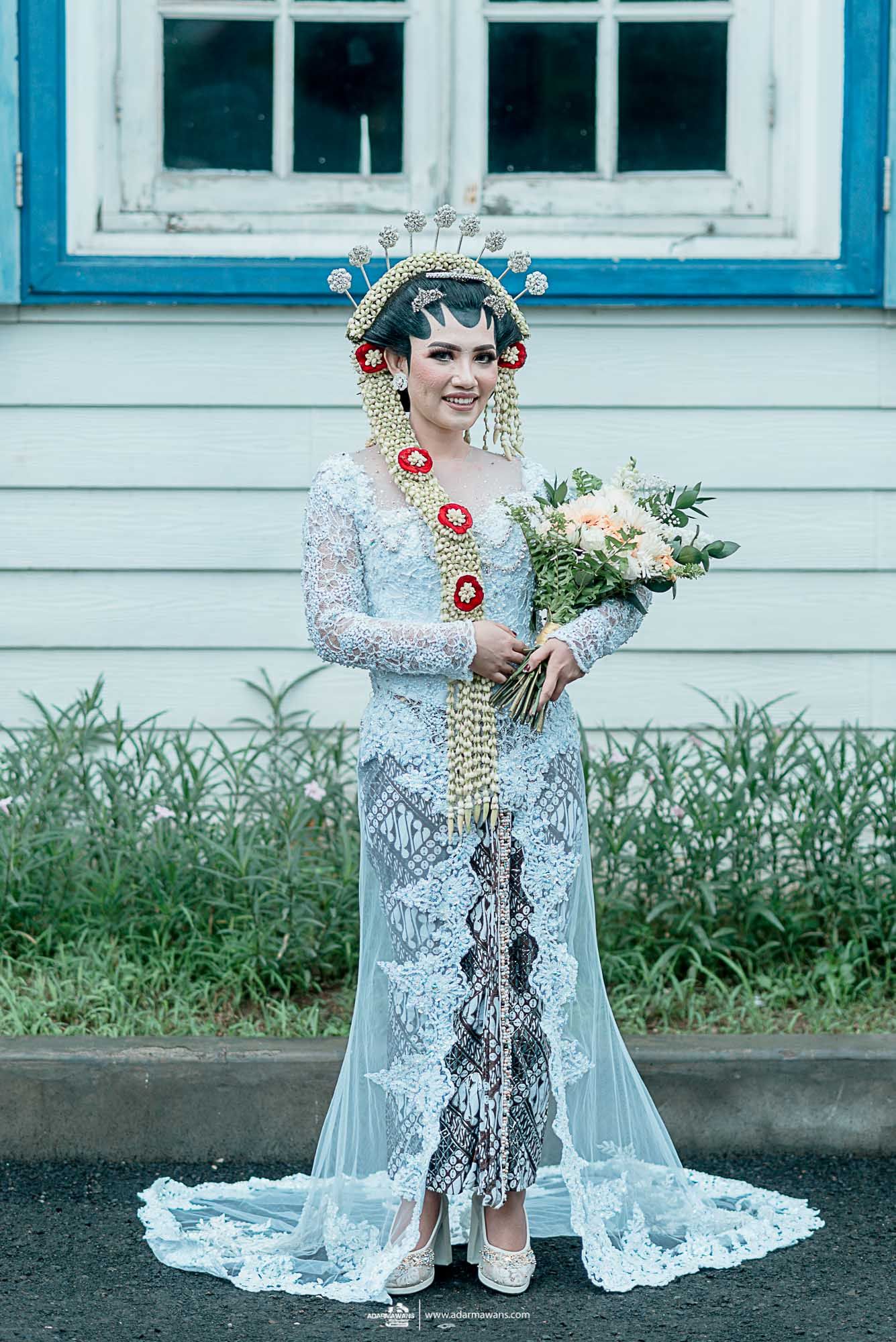 Bride in ornate lace wedding dress with floral headpiece holding bouquet against white building.