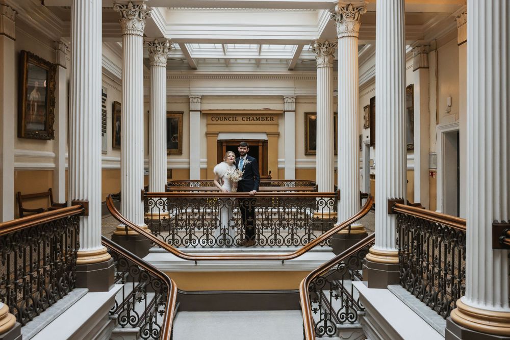 A bride and groom posing inside Brighton Town Hall, surrounded by elegant architecture with white columns