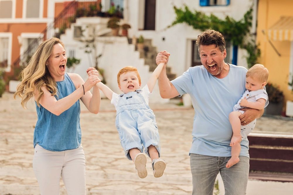Joyful parents playfully lifting toddler in overalls while holding baby in charming European setting.