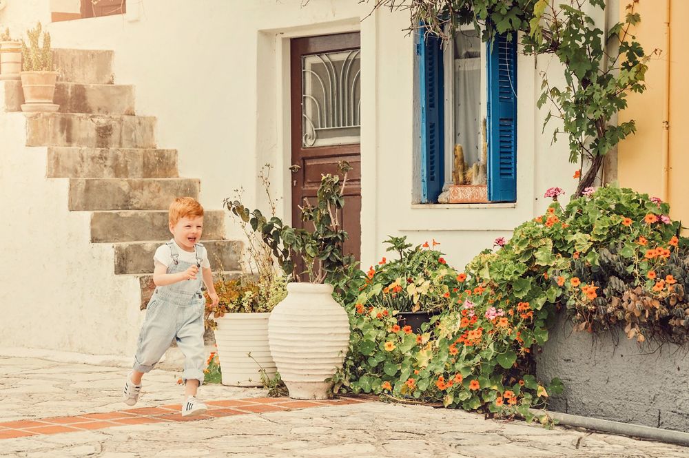 Young child in denim overalls running past white building with blue shutters and potted flowers.