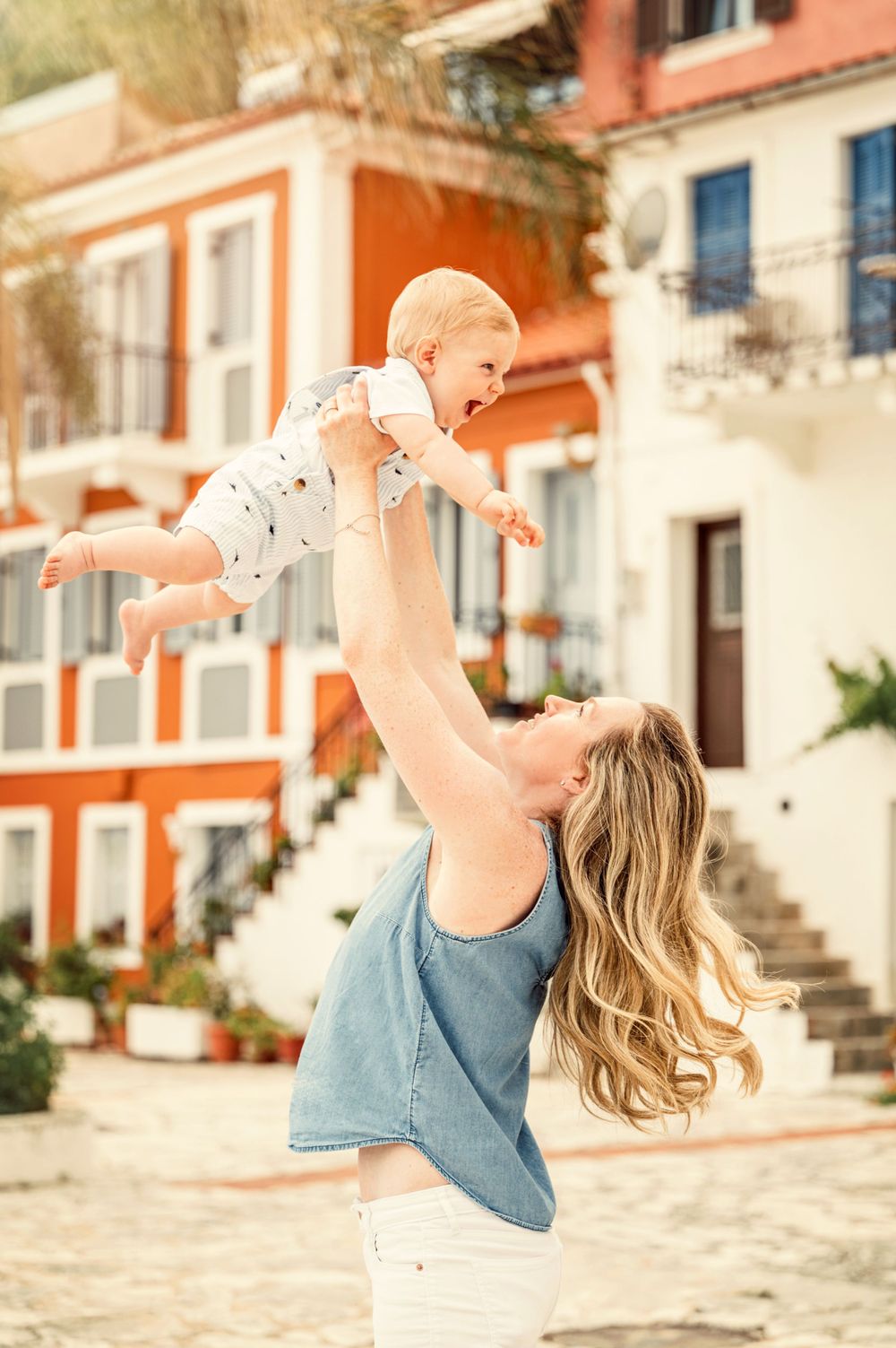 Mother lifting baby high in the air against backdrop of colorful European architecture.