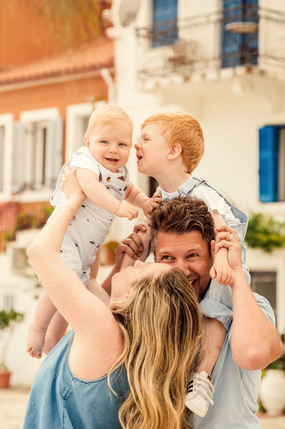 A joyful family moment with parents lifting their young children in front of a Mediterranean-style building.