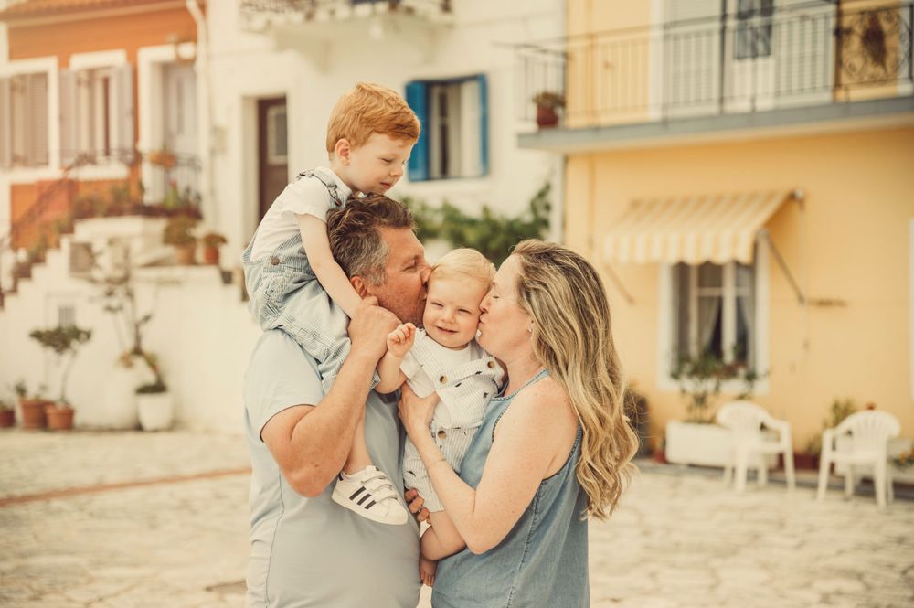 Candid family moment outdoors with parents holding children against pastel building backdrop.