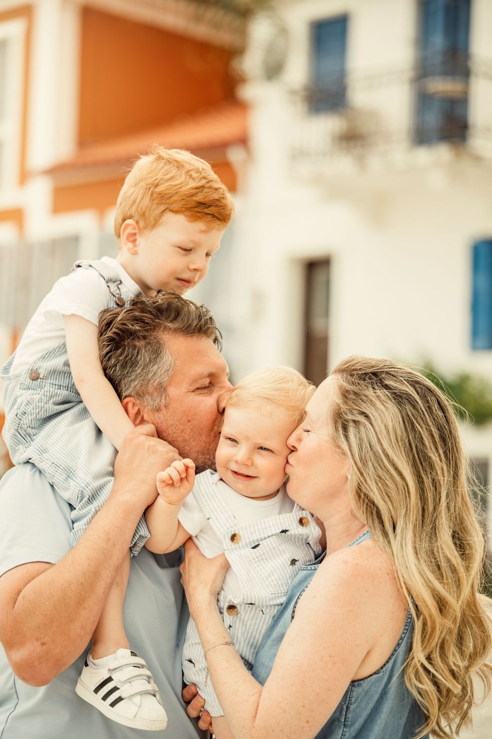Intimate family portrait with parents kissing baby while toddler rides on shoulders.