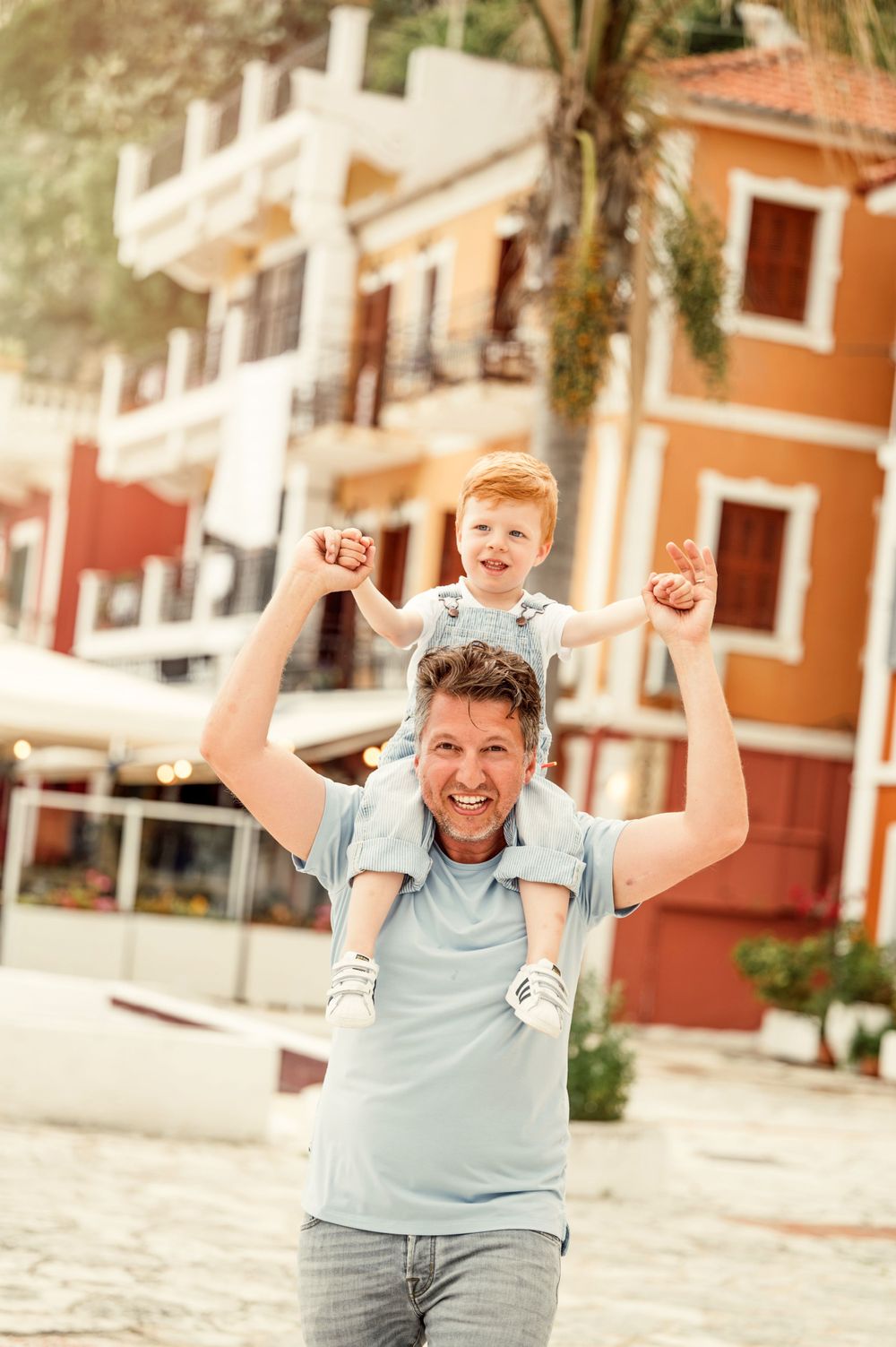 Father and child celebrating with arms raised against orange Mediterranean buildings.