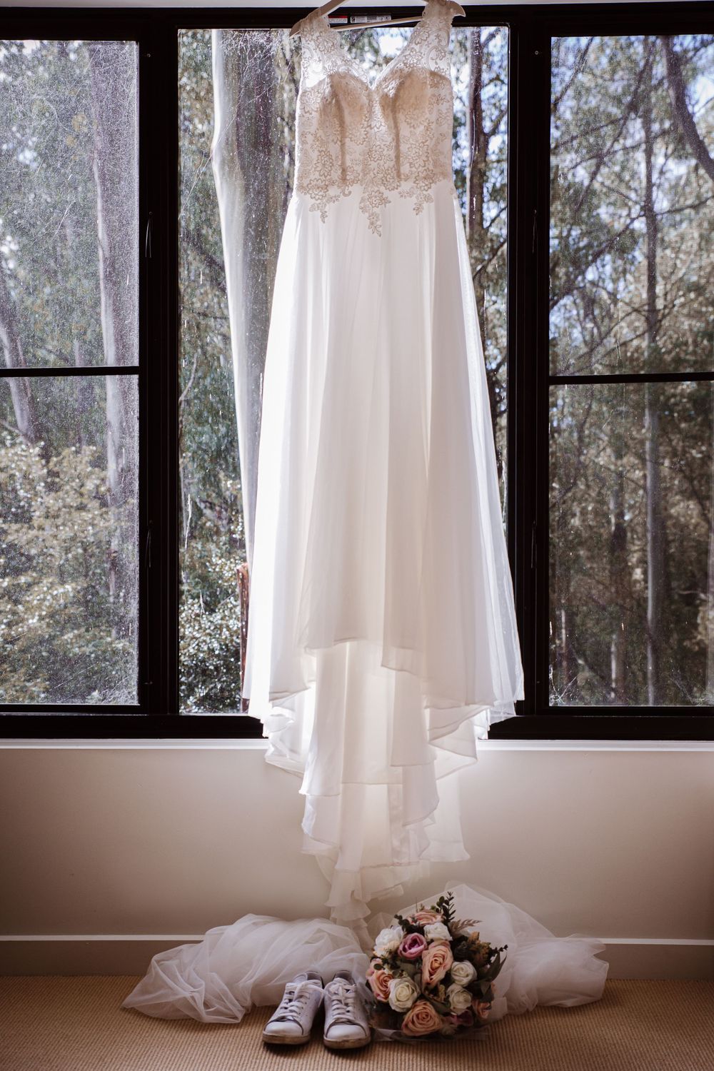 White wedding dress hanging in front of large windows with sunlight streaming through trees in background.