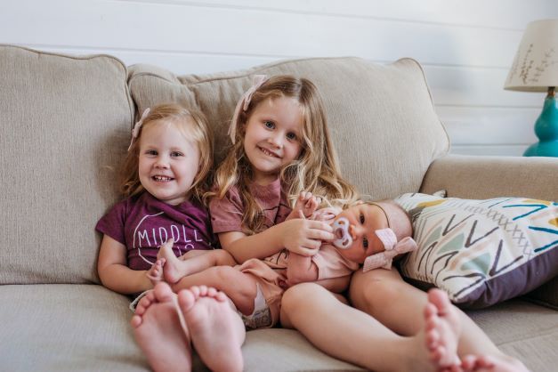 Two young girls hold new baby sister on a couch in their home