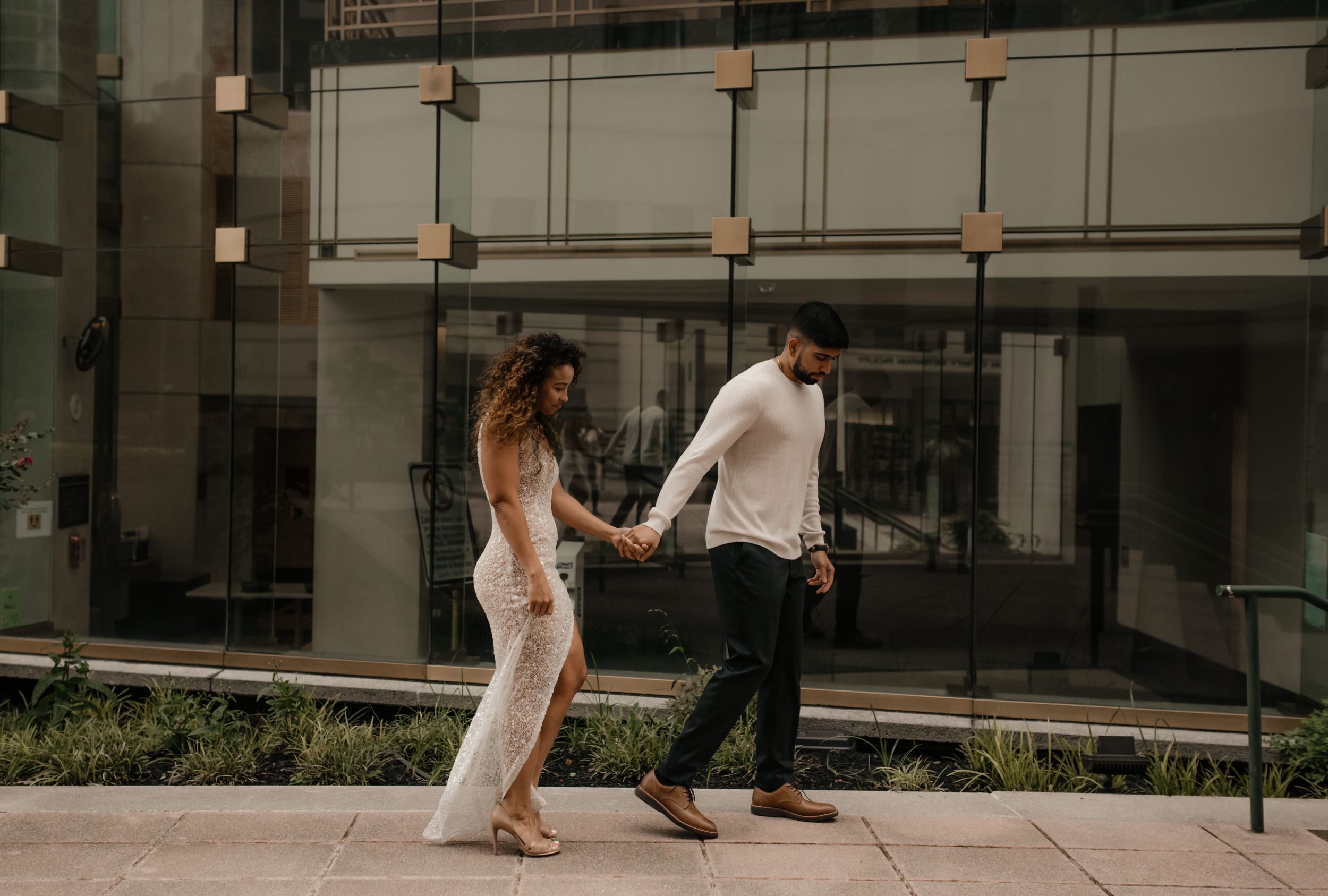 A couple holds hands while walking along a modern building exterior.