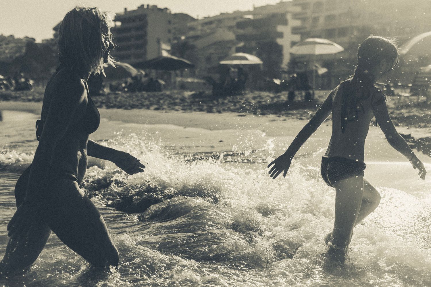 A mother and daughter run through the shallow waves at the beach, silhouetted against the late afternoon sun. The water splashes their feet.