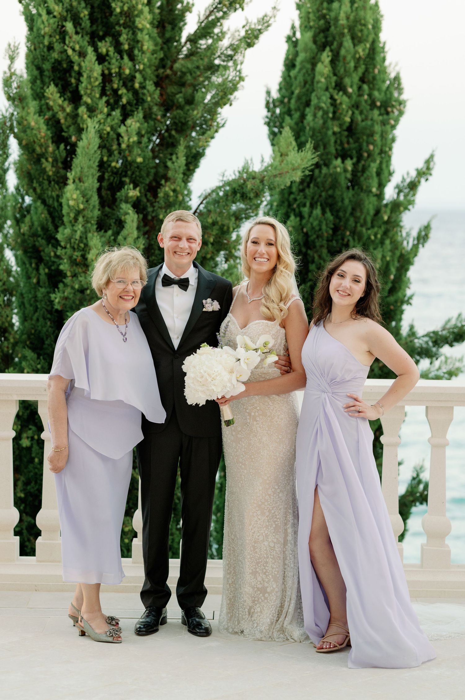 Wedding party poses against cypress trees on an elegant Mediterranean terrace overlooking the sea.
