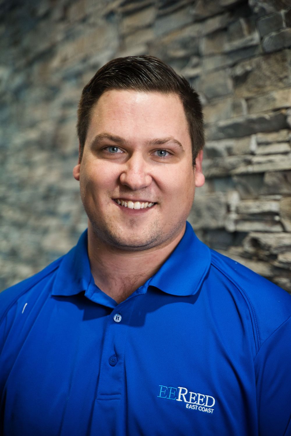 Professional headshot of a person in a blue EE Reed polo shirt smiling against a textured stone wall background.