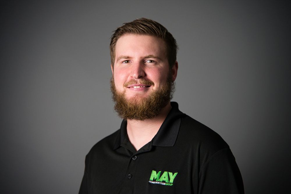 Professional headshot of a person wearing a black polo with May company logo against grey background.